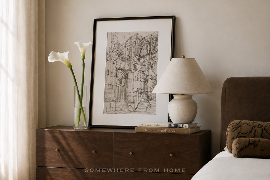 A framed black and white architectural sketch print displayed on a dark wood nightstand in a bedroom setting, alongside a vase with white calla lilies, a table lamp, and a bed.