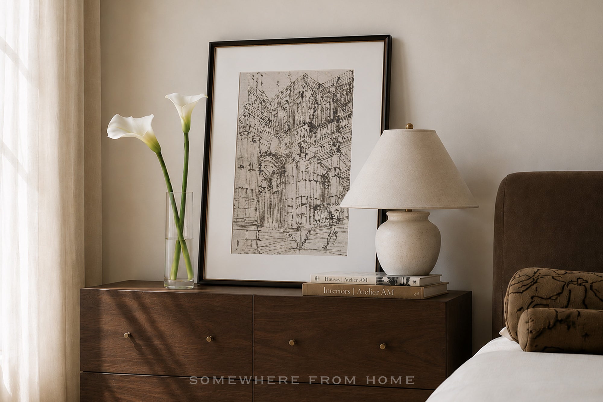 A framed black and white architectural sketch print displayed on a dark wood nightstand in a bedroom setting, alongside a vase with white calla lilies, a table lamp, and a bed.