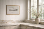 A framed black and white landscape print hangs on a white paneled wall above a marble countertop with a rustic ceramic vase of branches on the windowsill in a bright kitchen setting.