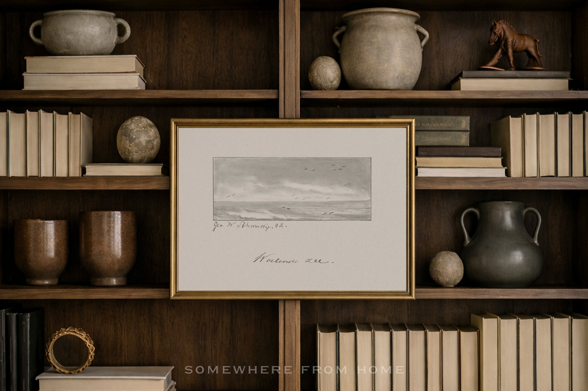 A framed black and white landscape sketch with elegant cursive writing, set in a gold frame and displayed on a dark wood bookshelf surrounded by books and decorative pottery.