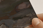 A close-up of a hand holding a square art print or card showing an abstract painting with thick brushstrokes in dark gray, brown, olive green, and off-white. The text 'SOMEWHERE FROM HOME' appears near the bottom.