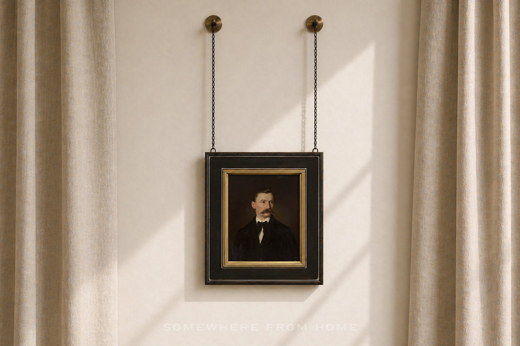 A framed vintage-style portrait print hanging from two black chains on a beige wall between beige curtains. The portrait shows a man in a dark suit with a white shirt and bow tie, set within a black frame with a gold inner border.