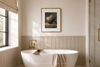 A styled bathroom interior featuring a white freestanding bathtub with a gold faucet, a beige towel draped over the tub edge, a framed still life print on the wall, and a window with a beige blind on the left.