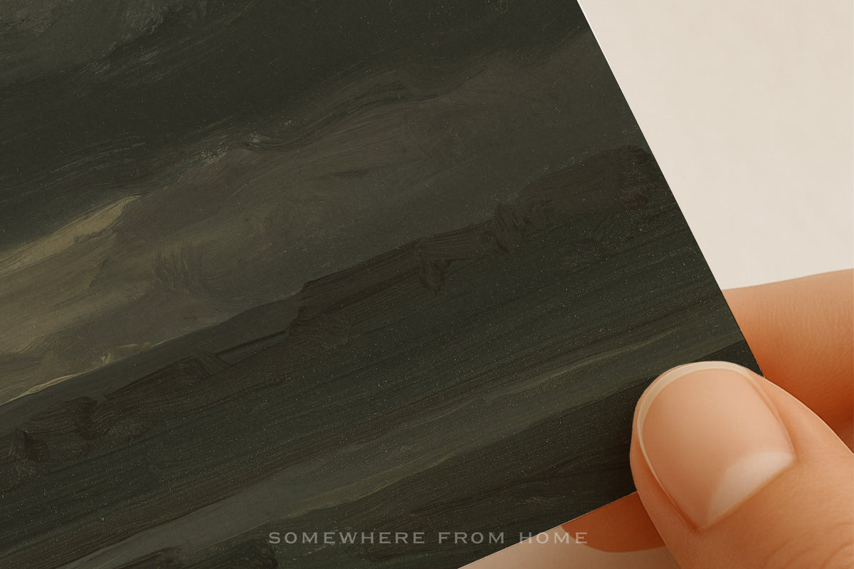 Hand holding a dark green landscape paper print on textured matte paper with 'SOMESWHERES FROM HOME' text.