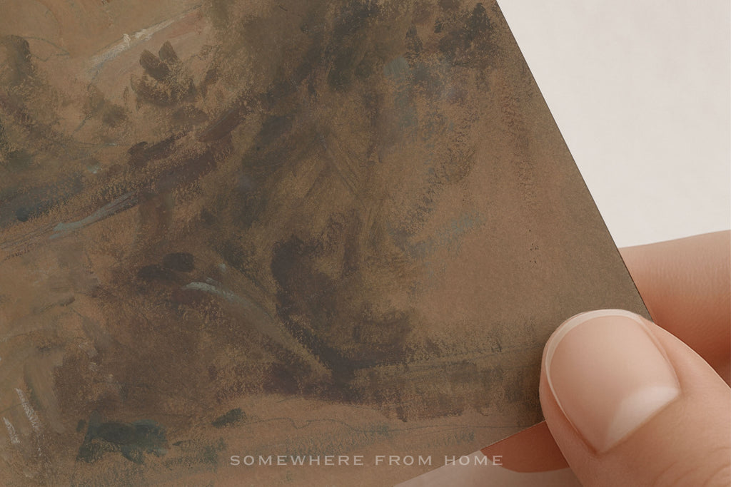 A close-up of a hand holding a textured art print or card featuring a muted landscape painting in earthy brown, gray, and beige tones. The text 'SOMEWHERE FROM HOME' is printed at the bottom.