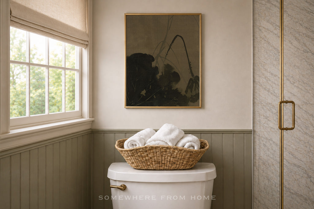 A framed black and white floral art print hangs on a beige wall above a toilet in a styled bathroom, with a woven basket of white towels resting on the toilet tank.
