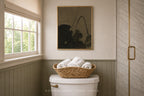 A framed black and white floral art print hangs on a beige wall above a toilet in a styled bathroom, with a woven basket of white towels resting on the toilet tank.
