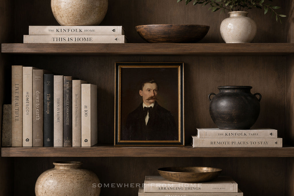 A framed vintage portrait print of a man with a mustache, displayed on a dark wooden shelf alongside books, ceramic vases, and bowls in a styled interior setting.