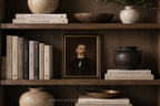 A framed vintage portrait print of a man with a mustache, displayed on a dark wooden shelf alongside books, ceramic vases, and bowls in a styled interior setting.