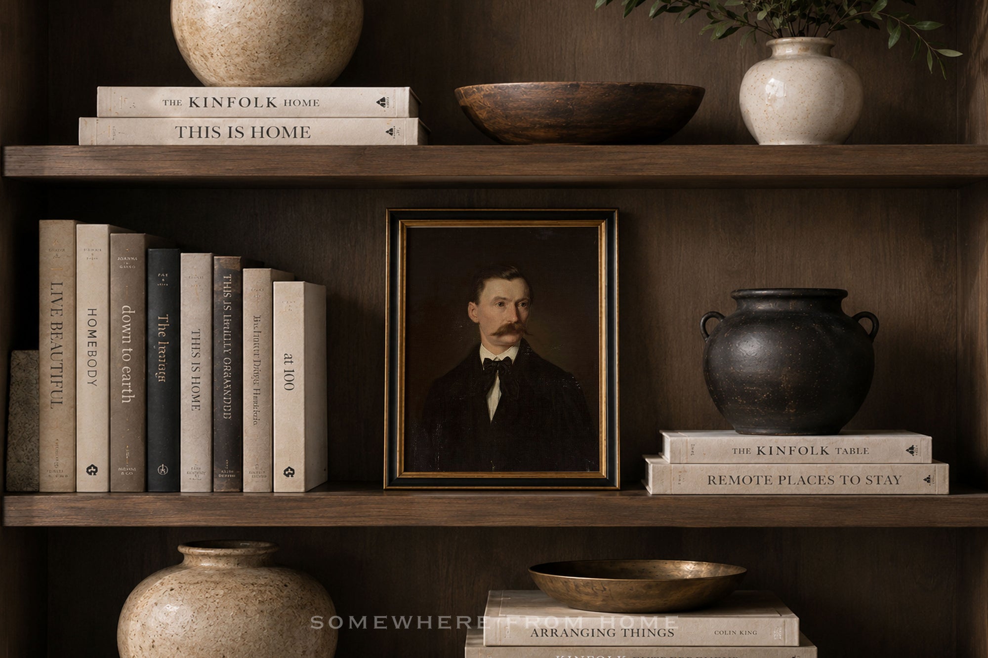 A framed vintage portrait print of a man with a mustache, displayed on a dark wooden shelf alongside books, ceramic vases, and bowls in a styled interior setting.