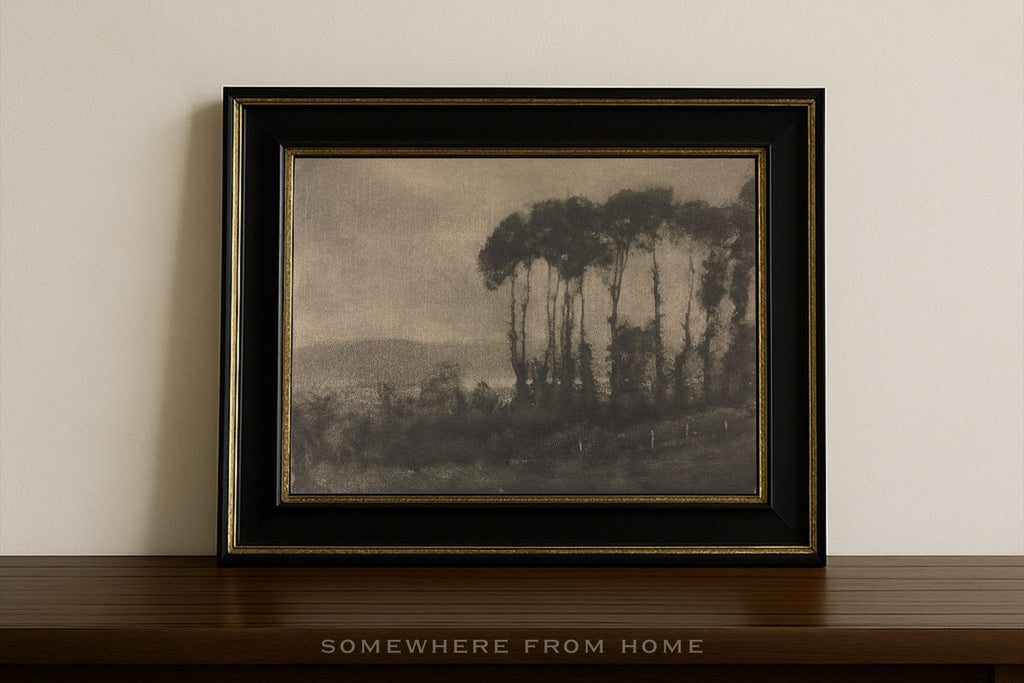 A framed landscape art print featuring a scene of tall trees and distant hills, presented in a black frame with gold trim on a dark wooden shelf against a light beige wall.