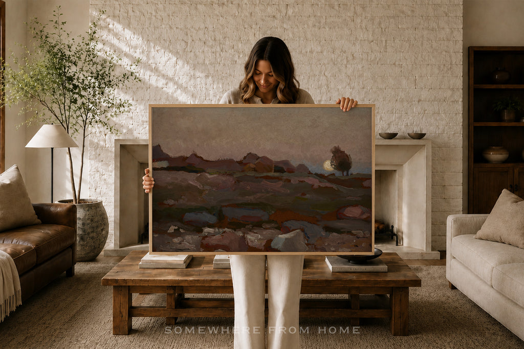 A woman stands in a cozy living room holding up a large framed landscape painting. The painting depicts a muted, earthy scene with hills, rocks, and a small tree.