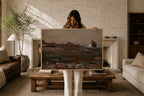 A woman stands in a cozy living room holding up a large framed landscape painting. The painting depicts a muted, earthy scene with hills, rocks, and a small tree.
