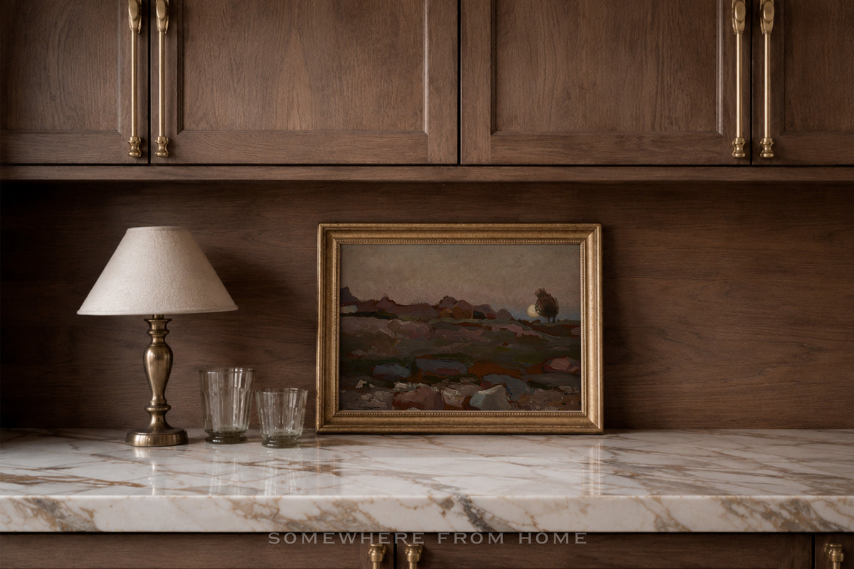 A framed landscape painting with a gold frame sits on a white marble countertop against a dark wood-paneled wall, next to a table lamp and two clear drinking glasses.