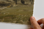 A close-up of a textured oil painting showing brushstrokes of olive green, brown, and black, held by a hand along its torn paper edge against a white background.
