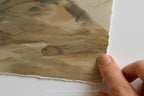A close-up of a hand holding the edge of a watercolor painting on textured paper, showing the painted landscape and the torn paper edge against a white background.