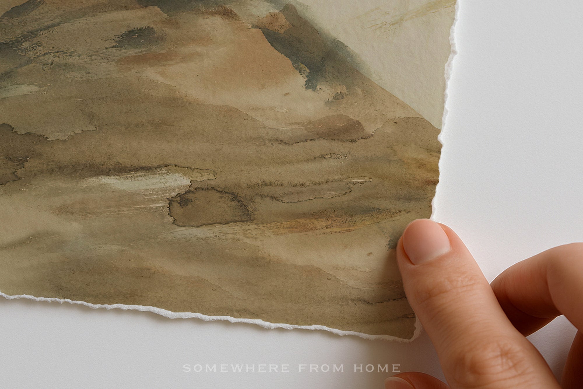A close-up of a hand holding the edge of a watercolor painting on textured paper, showing the painted landscape and the torn paper edge against a white background.