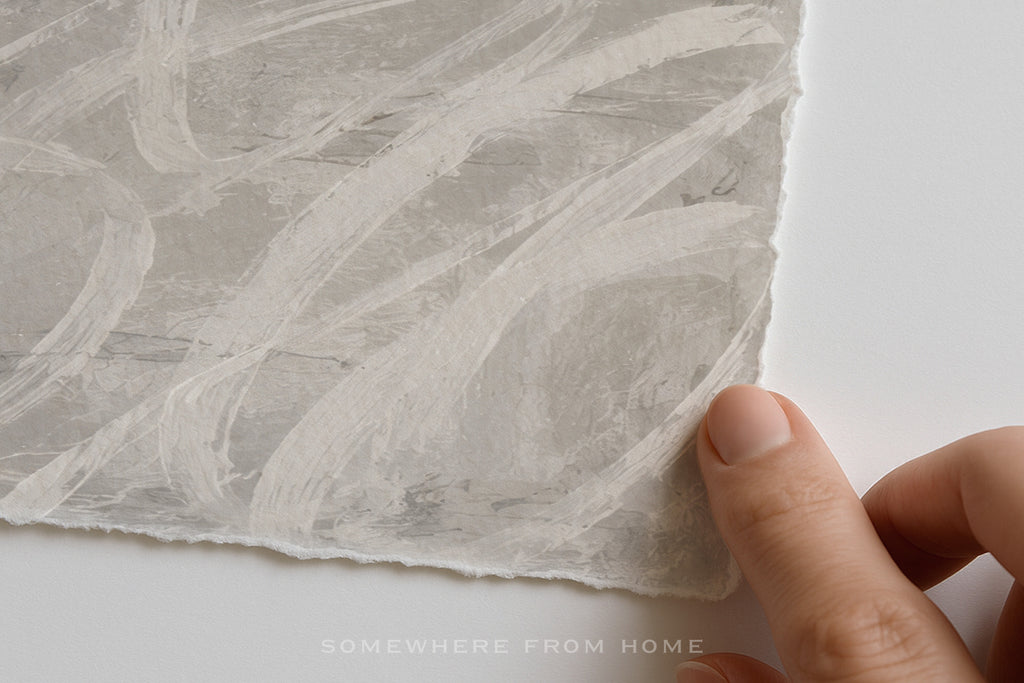 A close-up of a hand holding the edge of a square sheet of textured, handmade paper with a mottled off-white and light gray brushstroke pattern, set against a clean white background.