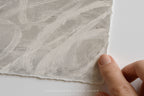 A close-up of a hand holding the edge of a square sheet of textured, handmade paper with a mottled off-white and light gray brushstroke pattern, set against a clean white background.