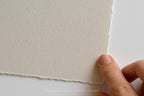 A close-up of a hand holding the edge of a square sheet of textured off-white paper against a clean white background. The paper has a rough, torn edge and a matte finish.
