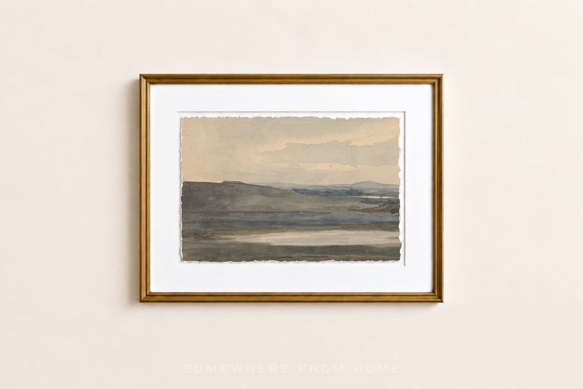 A framed landscape art print with a gold frame and white mat, hanging on a light beige wall. The artwork depicts a muted, minimalist scene of rolling hills under a pale sky.