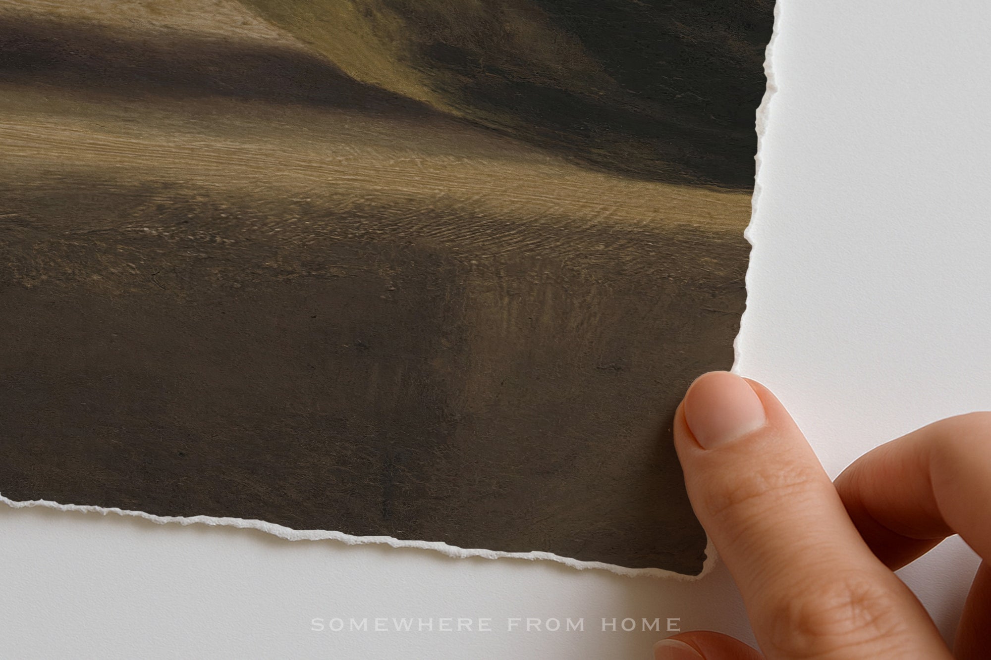 A close-up of a hand holding the edge of a torn paper print against a white background, showing a dark landscape artwork with earthy brown and olive tones. The text 'SOMEWHERE FROM HOME' appears at the bottom.