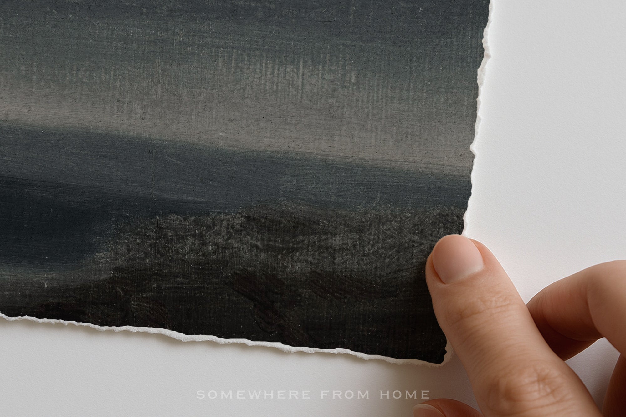 A close-up of a hand holding the edge of a torn sheet of textured paper with a dark charcoal landscape painting on it, set against a clean white background.