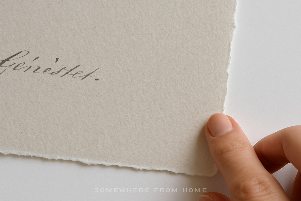A close-up of a hand holding the edge of a textured off-white paper card with torn margins, showing part of a handwritten script on the left side against a clean white background.