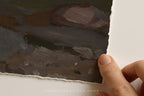 A close-up of a hand holding the edge of a textured, hand-painted art print or canvas with a torn paper look, showing various earthy brushstrokes of brown, gray, and green. A faint watermark at the bottom reads 'SOMEWHERE FROM HOME'.