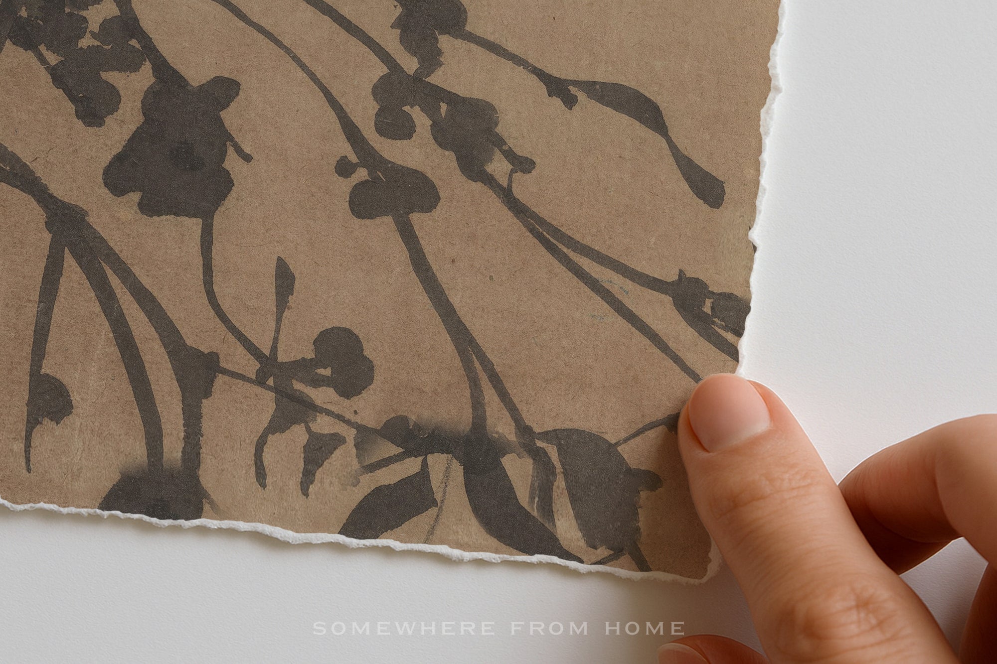 A close-up of a hand holding the edge of a square piece of textured brown paper featuring a dark ink floral branch pattern, against a plain white background.