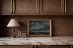 A framed landscape painting of a stormy sea under a dark cloudy sky, displayed on a white marble countertop in a kitchen setting with dark wood cabinets, a table lamp, and two glasses.