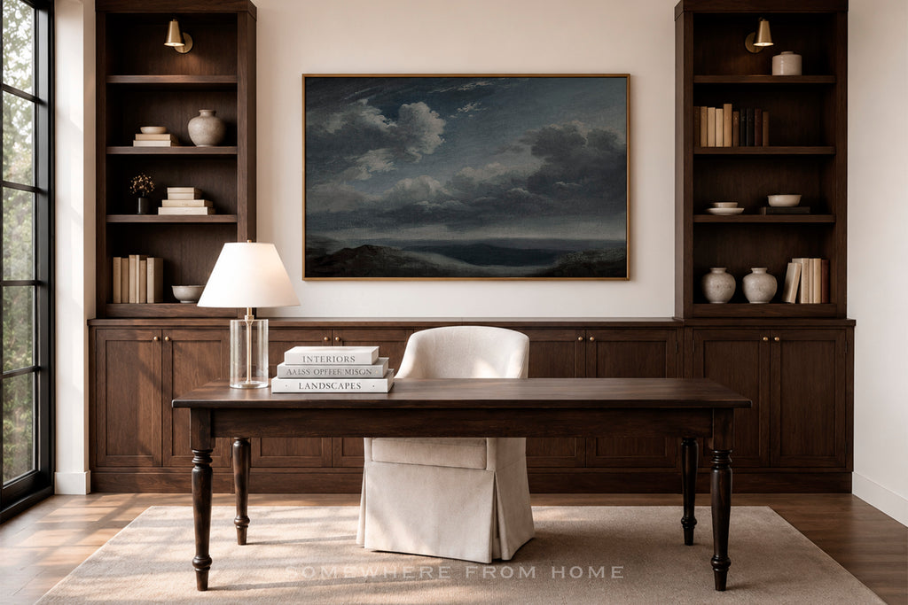 A large framed landscape painting of a stormy sea hangs on a beige wall in a home office setting, flanked by dark wood built-in shelves with books and vases. Below the painting is a dark wood desk with a white chair slipcover, a table lamp, and stacked books.