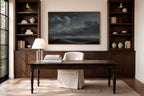 A large framed landscape painting of a stormy sea hangs on a beige wall in a home office setting, flanked by dark wood built-in shelves with books and vases. Below the painting is a dark wood desk with a white chair slipcover, a table lamp, and stacked books.