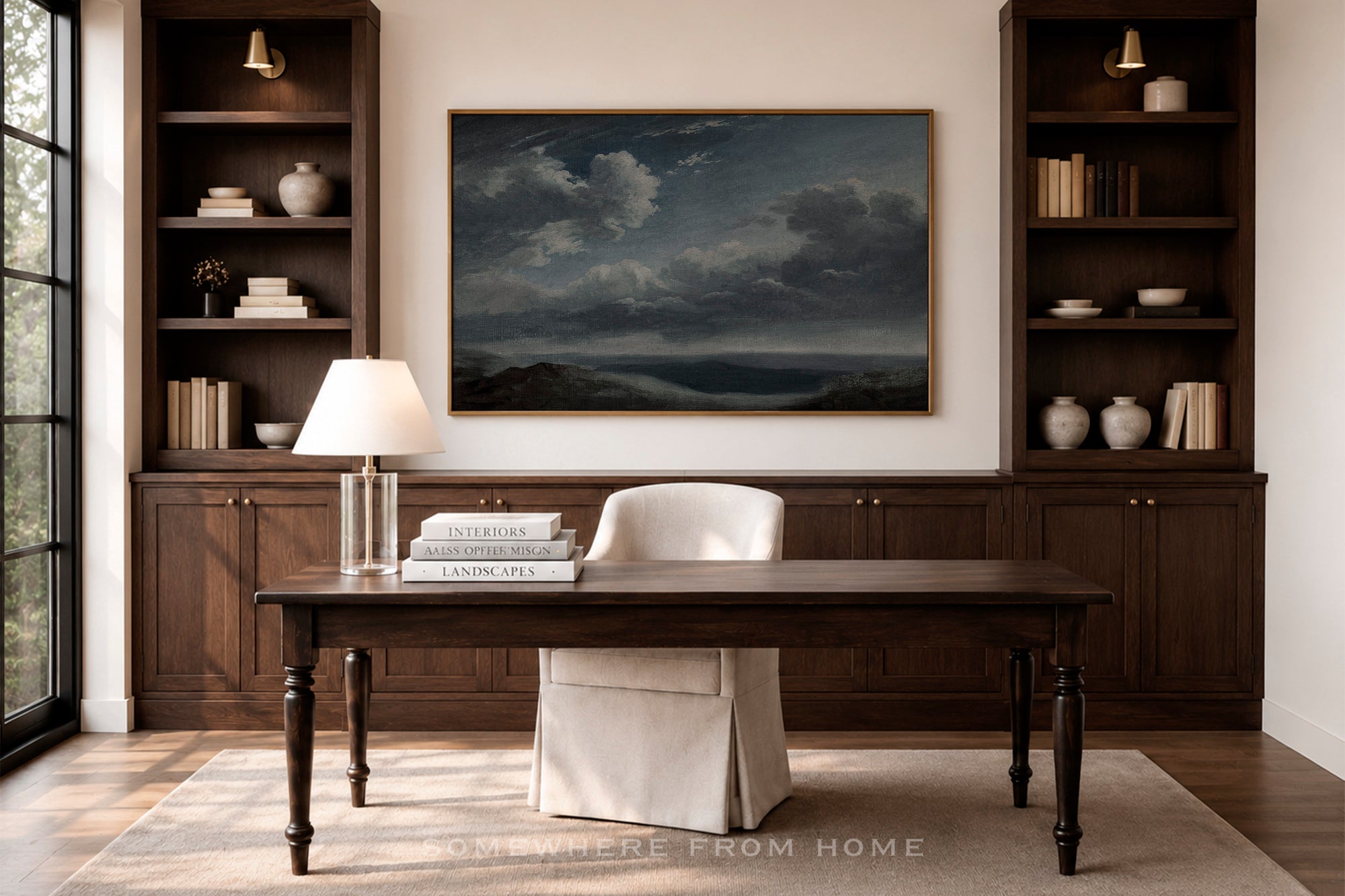 A large framed landscape painting of a stormy sea hangs on a beige wall in a home office setting, flanked by dark wood built-in shelves with books and vases. Below the painting is a dark wood desk with a white chair slipcover, a table lamp, and stacked books.