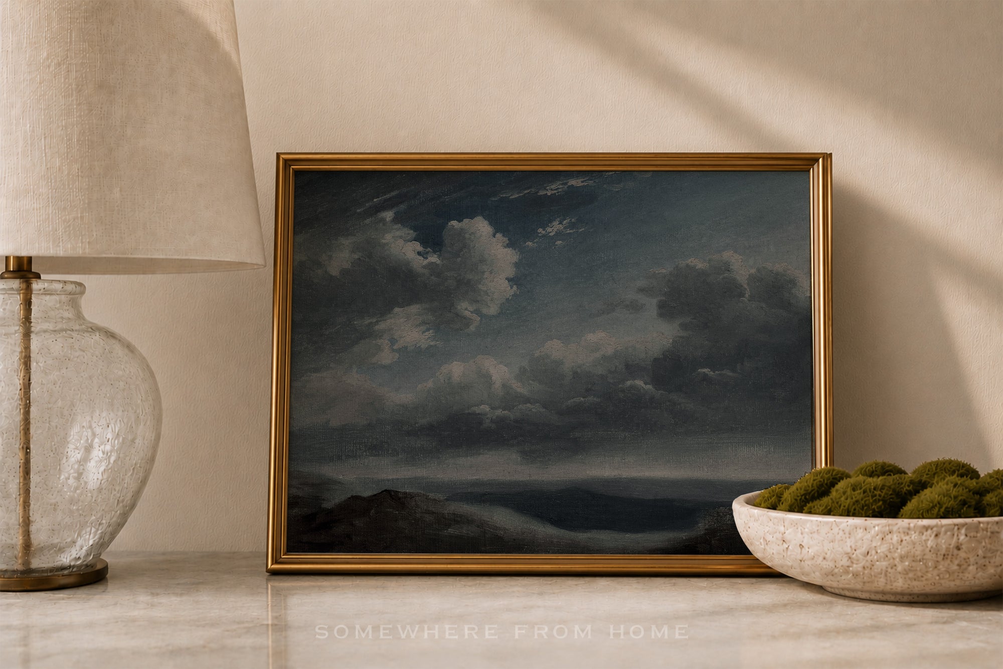 A framed landscape painting of a stormy sky over dark mountains, styled on a marble surface next to a lamp and a bowl of green moss.
