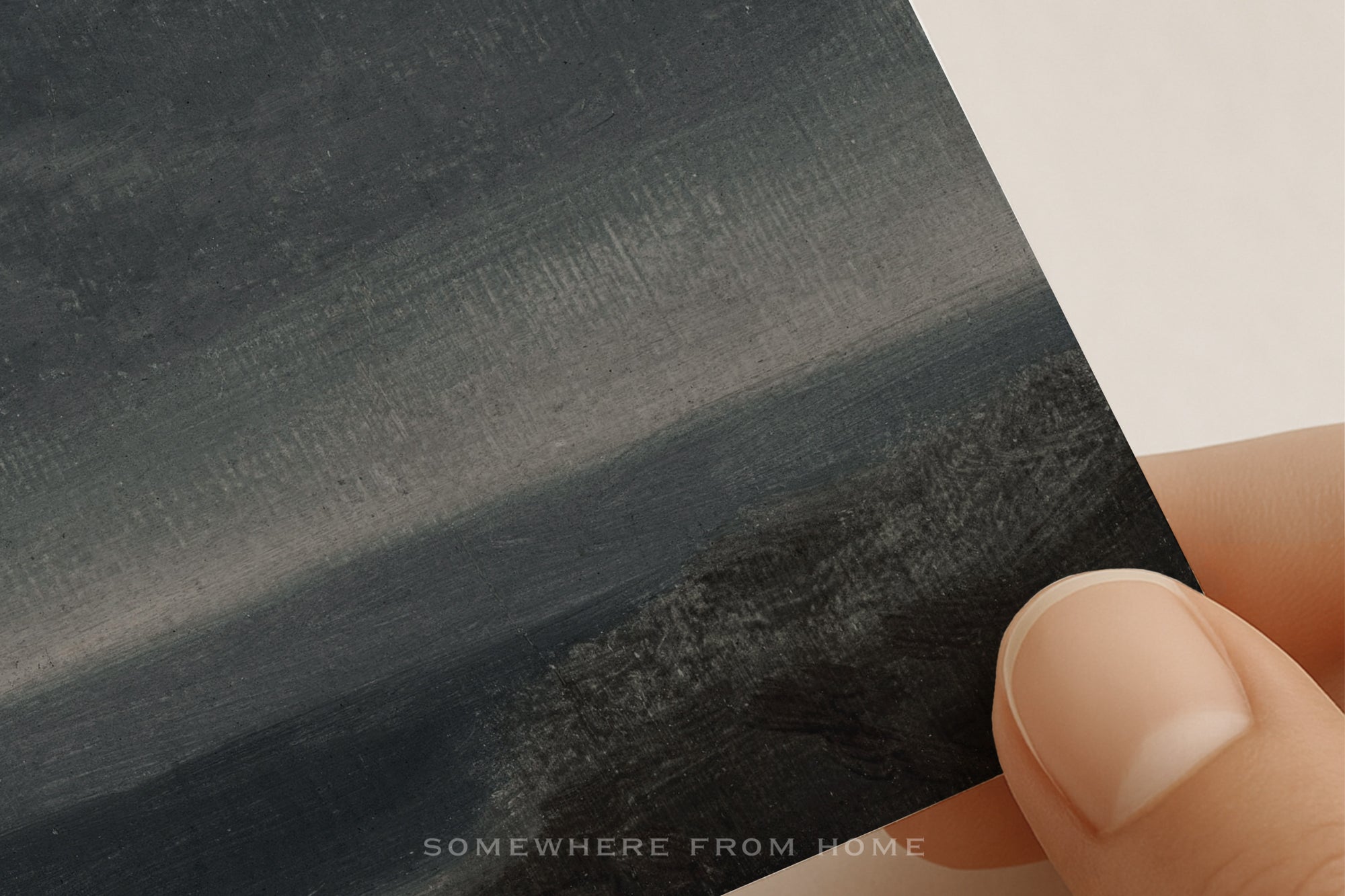 A close-up of a hand holding a textured art print or card featuring a dark, painterly landscape in black, gray, and charcoal tones against a white background.