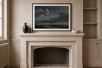 A framed landscape art print depicting a stormy sea under a dark cloudy sky, displayed on a beige stone fireplace mantel in a classic room setting.