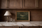 A framed landscape painting of a stormy sky hangs on a dark wood wall above a marble countertop with a table lamp and two glasses.