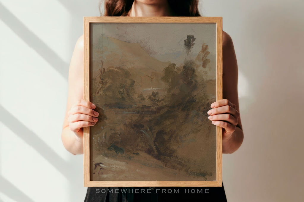 A person holds a framed landscape painting with a light wood frame against a white wall with soft diagonal shadows. The painting depicts a muted, earthy scene in brown, beige, and gray tones.