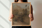 A person holds a framed landscape painting with a light wood frame against a white wall with soft diagonal shadows. The painting depicts a muted, earthy scene in brown, beige, and gray tones.