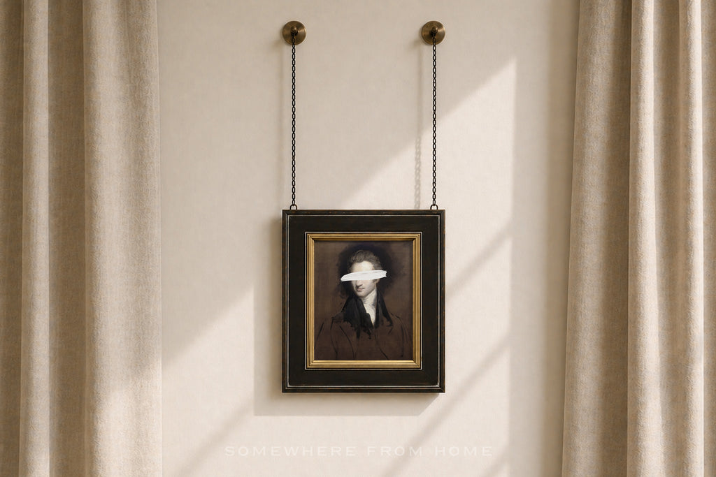 A framed portrait print hanging from two chains on a beige wall between sheer curtains. The artwork shows a dark-haired figure in a dark brown and black outfit with a white blindfold over the eyes, set in a black frame with a gold inner border.