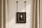 A framed portrait print hanging from two chains on a beige wall between sheer curtains. The artwork shows a dark-haired figure in a dark brown and black outfit with a white blindfold over the eyes, set in a black frame with a gold inner border.