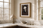 A framed portrait print hangs on a cream wall above a marble countertop in a sunlit room. The artwork shows a dark-haired woman with a white blindfold over her eyes, enclosed in a gold frame.