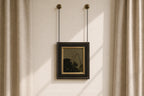 A framed art print with a black frame and gold inner border hangs from two black chains on a beige wall between beige curtains.