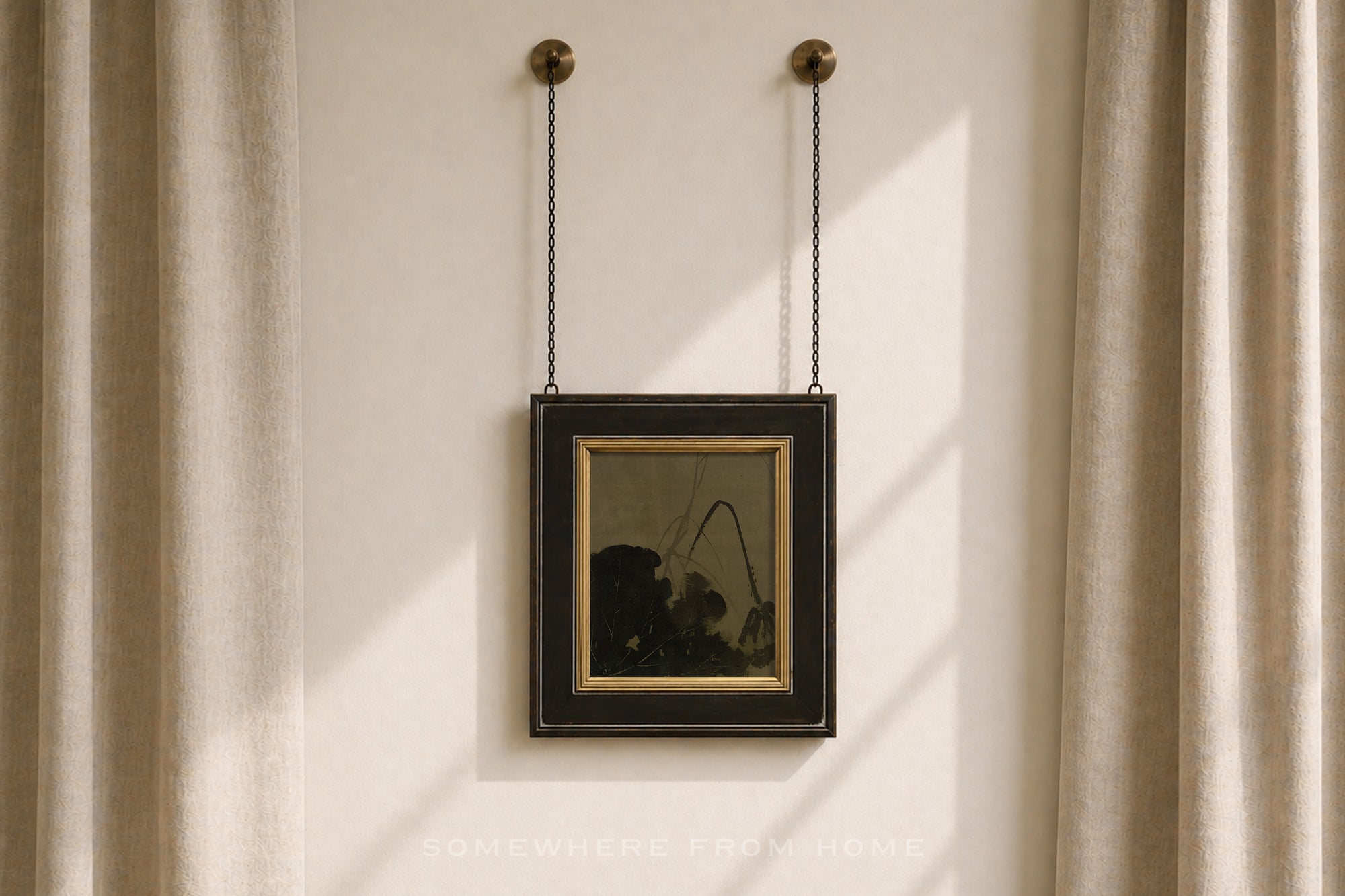 A framed art print with a black frame and gold inner border hangs from two black chains on a beige wall between beige curtains.