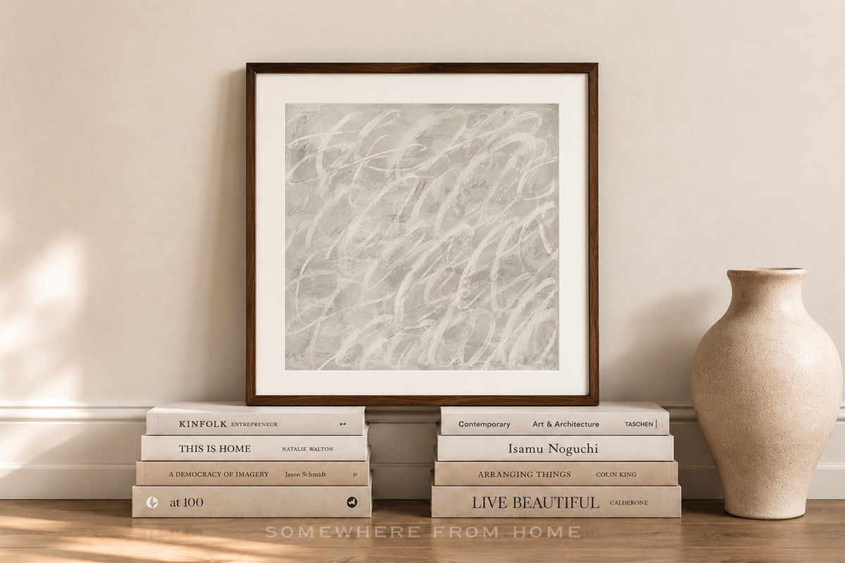 A framed abstract art print with a white mat and dark brown frame, leaning against a beige wall on a wooden floor, styled with books and a ceramic vase.