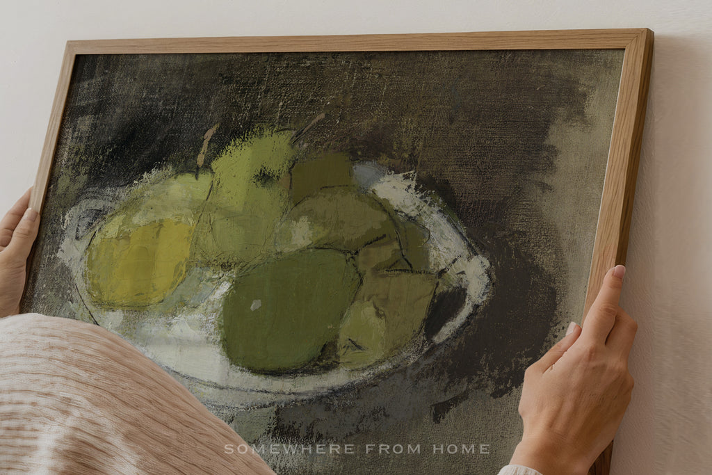 A framed still life painting of green pears on a dark textured background, held by a person whose hands and shoulder are visible against a light wall.