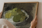 A framed still life painting of green pears on a dark textured background, held by a person whose hands and shoulder are visible against a light wall.