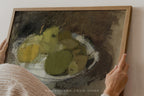 A framed still life painting of green and yellow pears on a dark brown background, held by a person whose hands and shoulder are visible against a light wall.