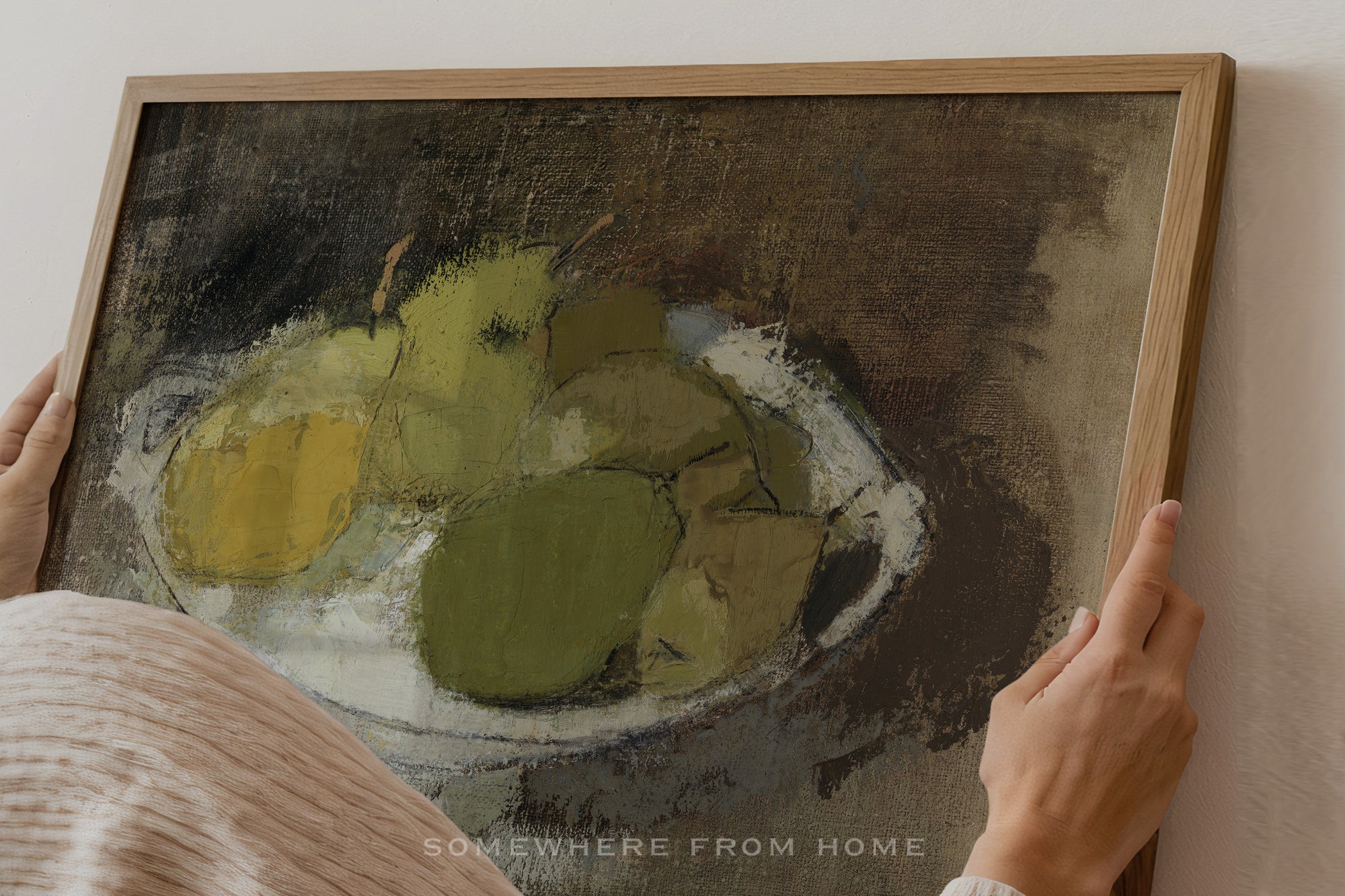 A framed still life painting of green and yellow pears on a dark brown background, held by a person whose hands and shoulder are visible against a light wall.